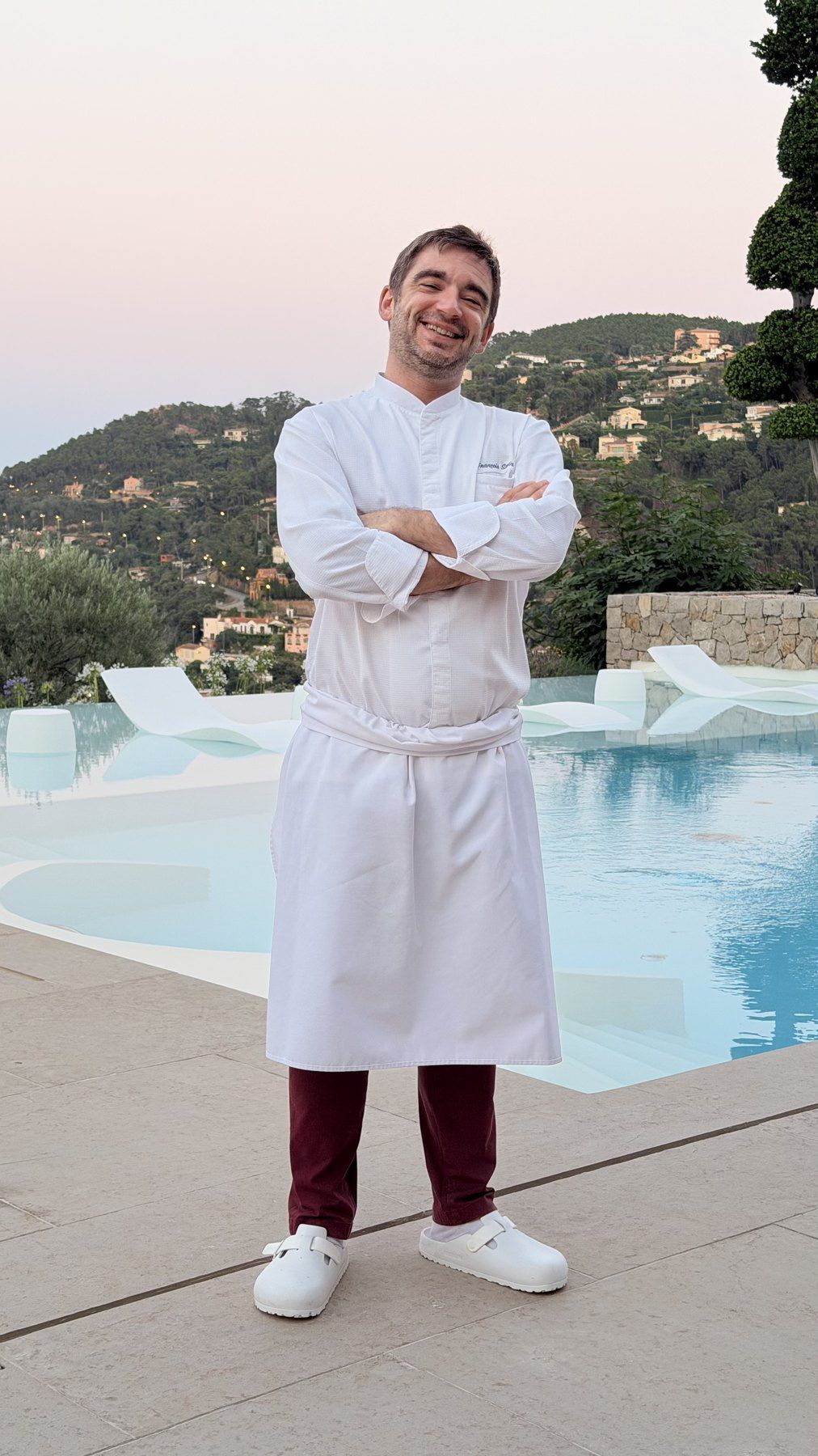 Chef François Savin in his whites and apron, arms crossed beside an infinity pool at dusk, wooded hills behind.
