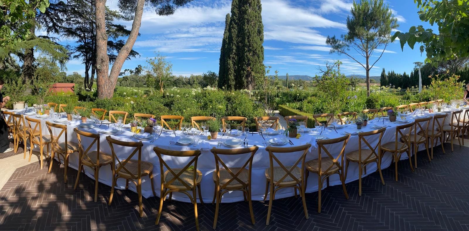 Long banquet table set with white linen and wooden bistro chairs in a Provençal garden, cypress and pines under a blue sky
