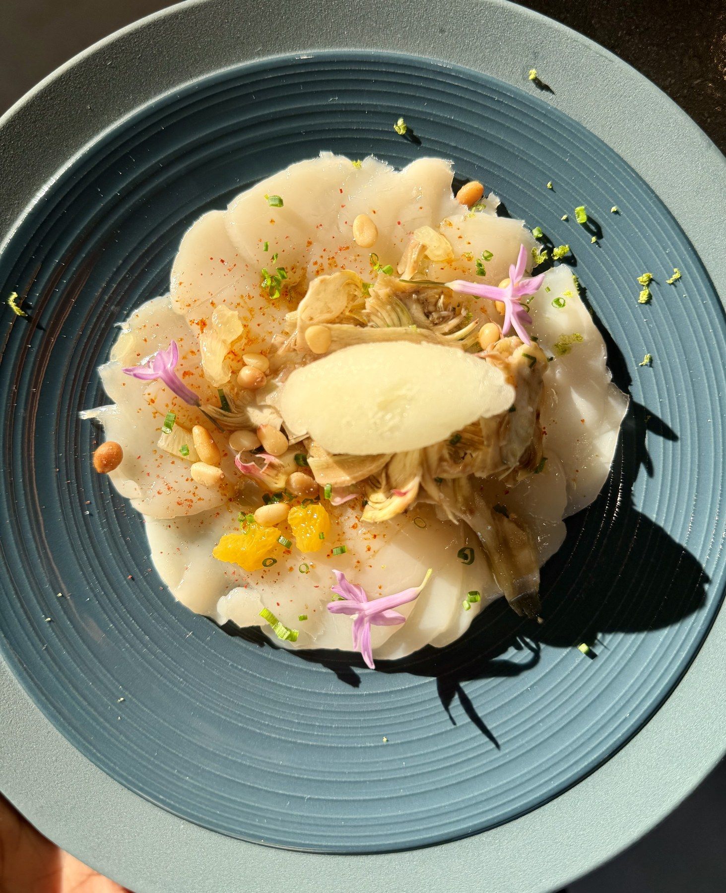 Carpaccio de Saint-Jacques à l’artichaut, pignons et fleurs comestibles sur assiette bleue.