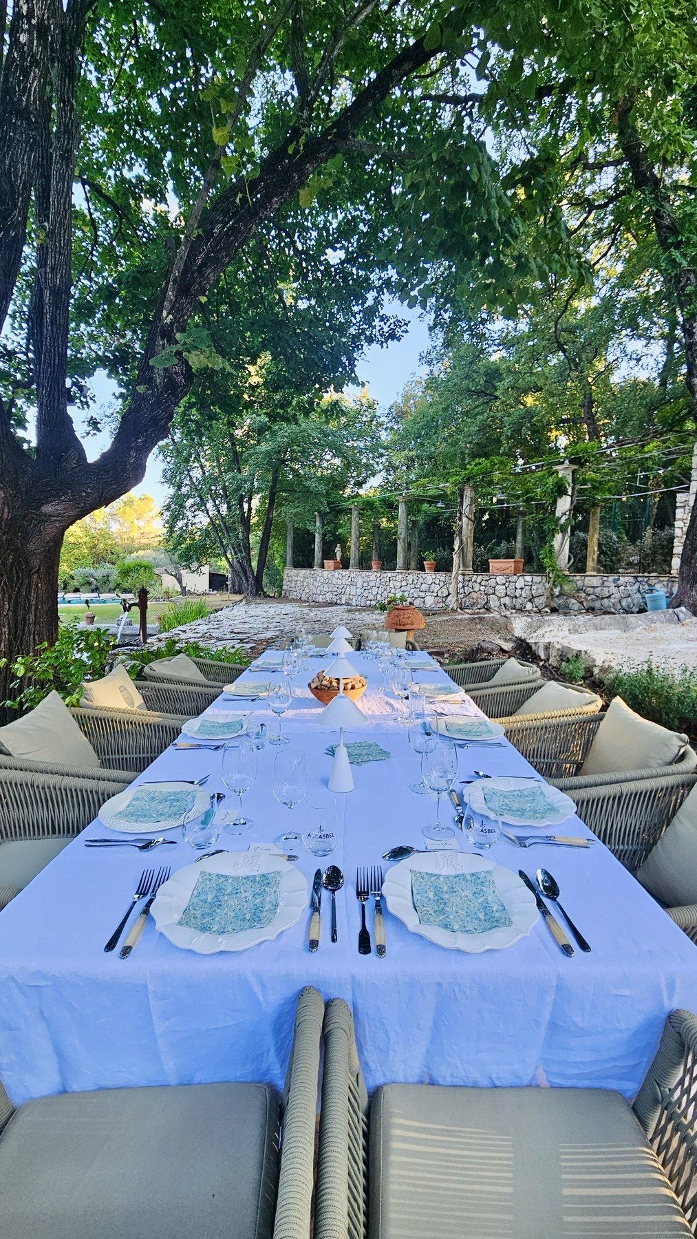 Longue table en lin bleu pâle dressée sous les arbres.