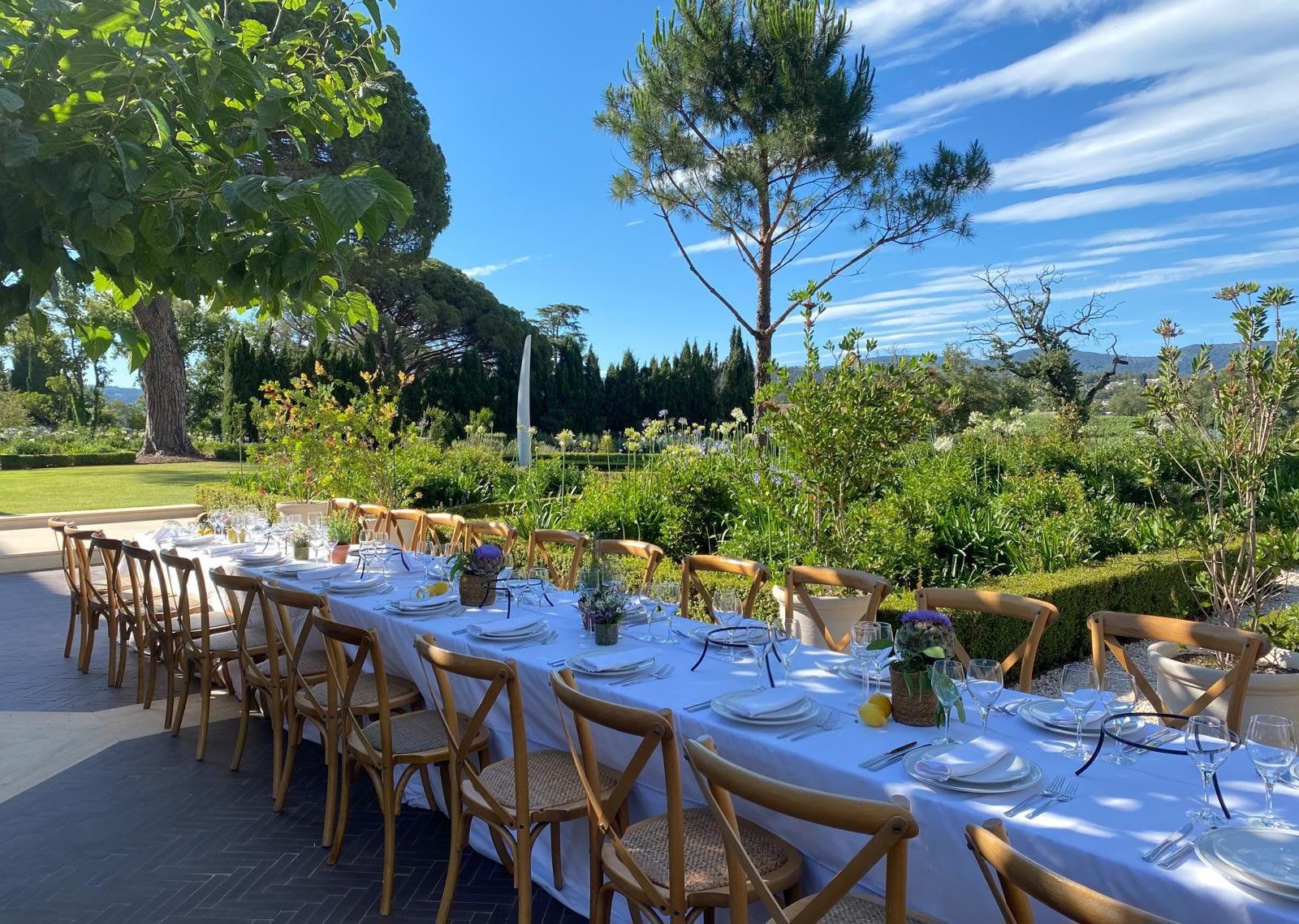 Long banquet table dressed in white linen in a Provençal garden, pines and hills beyond.