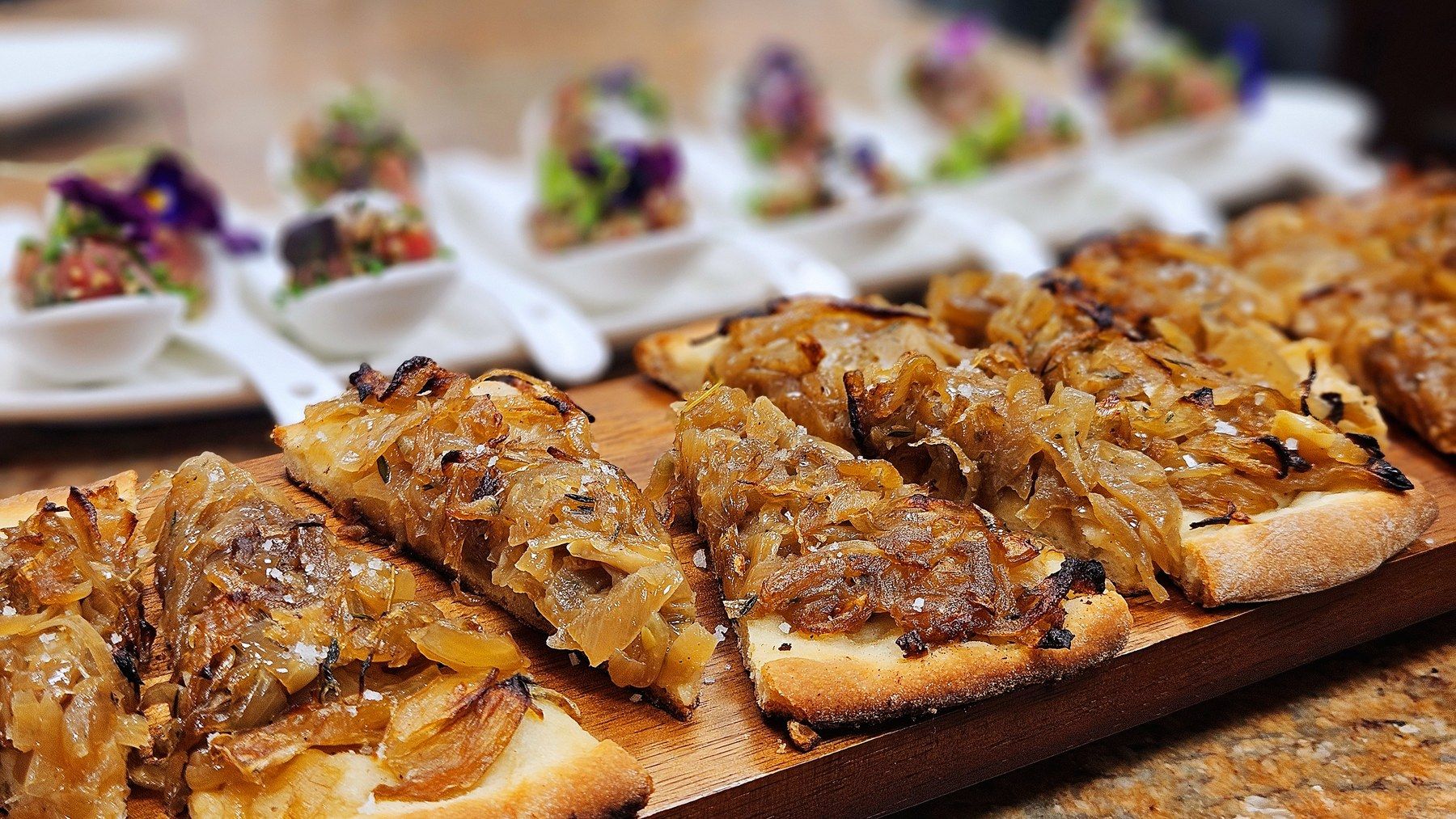Tartines d’oignons confits sur planche, cuillères d’amuse-bouche derrière.