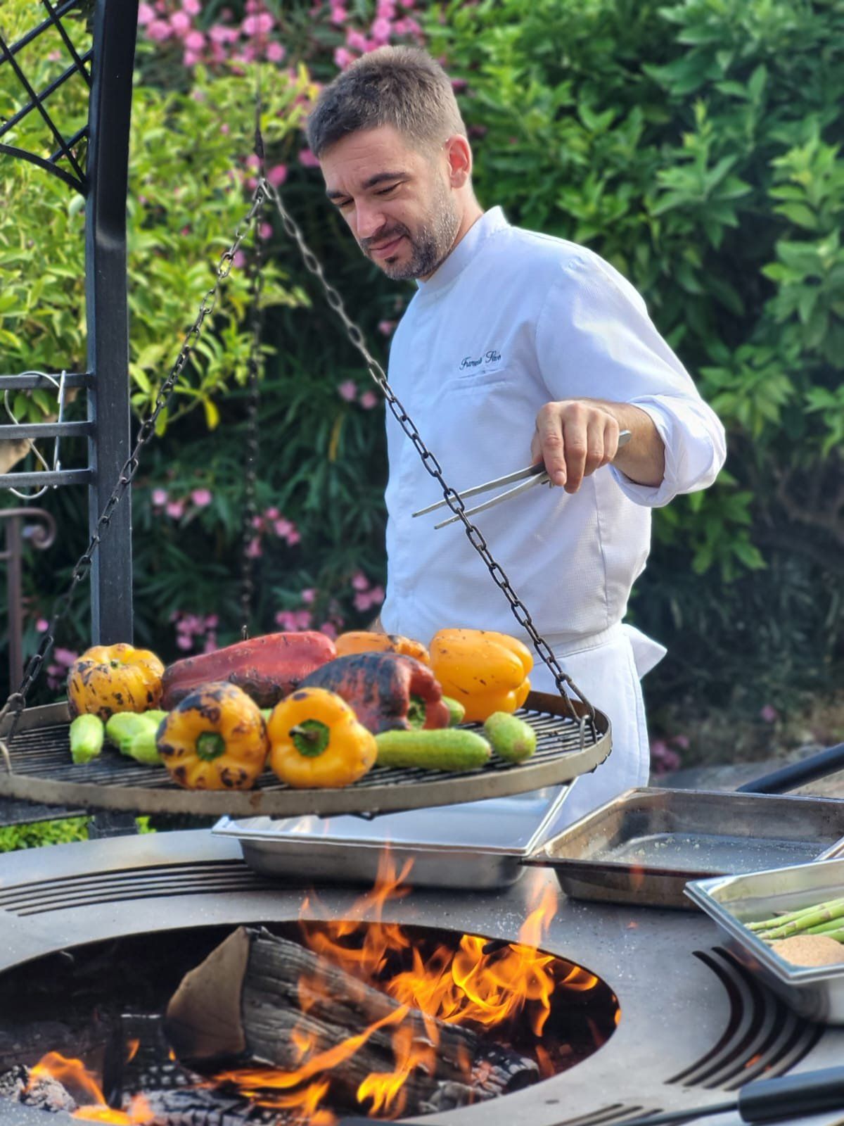 Chef in his whites grilling vegetables on a hanging plate above an open fire.